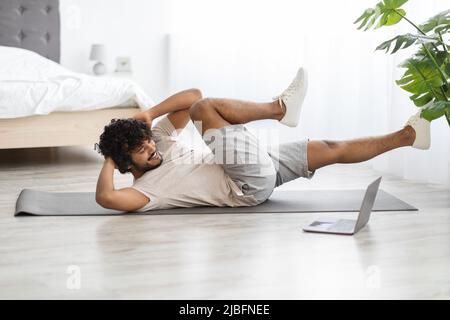 Aktiver Ostmann in Sportbekleidung, der zu Hause mit einem Laptop trainiert Stockfoto