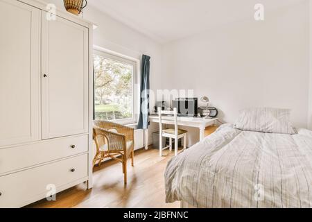 Korbsessel zwischen Schrank und Tisch mit Computer neben einem bequemen Bett im sonnendurchfluteten Schlafzimmer mit Fenster Stockfoto