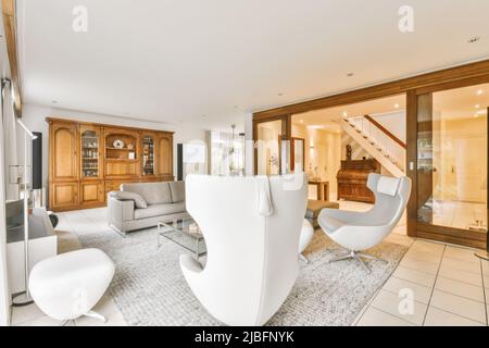 Komfortables Sofa mit Sesseln und Couchtisch aus Glas auf grauem Teppich im geräumigen, hellen Wohnzimmer mit Vintage-Holzschrank und Topfpflanzen Stockfoto