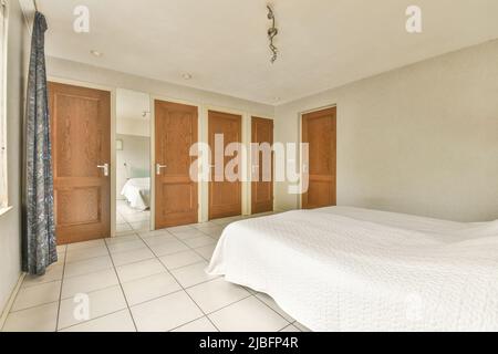 Komfortables Bett mit Bettdecke im hellen Schlafzimmer mit Fliesenboden und Holztüren in Reihe an sonnigen Tag im Haus Stockfoto