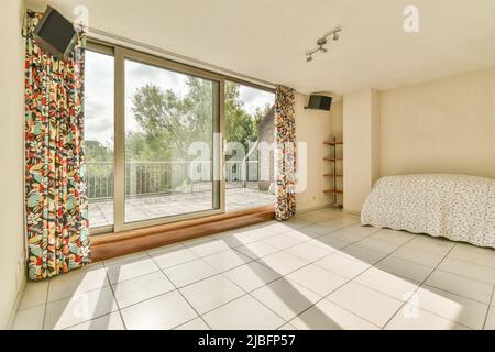 Komfortables Bett mit Bettdecke im hellen Schlafzimmer mit gefliestem Boden und Holztür mit Glastüren, die an sonnigen Tagen zum Balkon führen Stockfoto