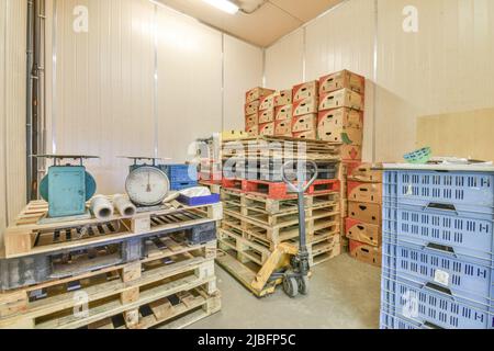 Alte Metallwaagen und gerollte Kunststoffverpackungen, die auf Holzpaletten in einem modernen Lagerhaus mit Kartons und Kunststoffbehältern gelagert werden Stockfoto