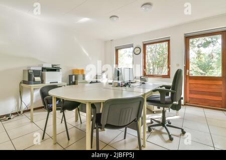 Tische mit modernen Computern und Druckern in einem geräumigen Büro mit Stühlen und gefliestem Boden in Sonnenlicht Stockfoto