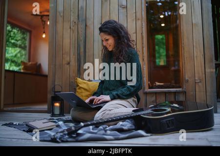 Glückliche junge Frau mit Laptop, die sich im Freien in einem Baumhaus, Wochenendausflug und Remote-Office-Konzept ausruhen lässt Stockfoto