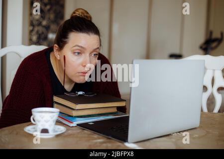 Konzentrierte müde Frau, die Informationen liest, Kaffee trinkt, Laptop benutzt, Fernarbeit zu Hause. Arbeitsprobleme im freiberuflichen Büro Stockfoto