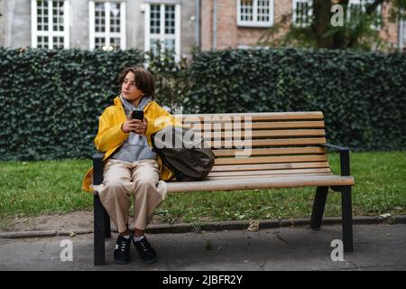 Das Mädchen sitzt auf der Bank und benutzt das Smartphone im Freien in der Stadt. Jemand wartet. Stockfoto