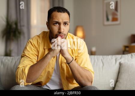 Schwarzer Mann, der mit nachdenklichem Gesichtsausdruck auf der Couch sitzt, sich traurig fühlt, Probleme hat, zu Hause unter Stress leidet Stockfoto