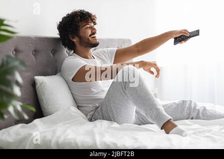 Fröhlicher inder, der auf dem Bett sitzt und fernsieht Stockfoto