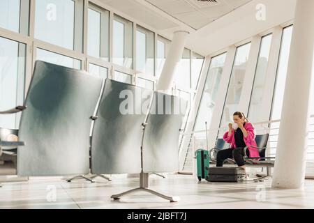 Porträt einer Frau, die eine weiße Einwegmaske aufsetzt und auf einem Stuhl neben Koffern vor einer grauen Sitzreihe sitzt. Stockfoto