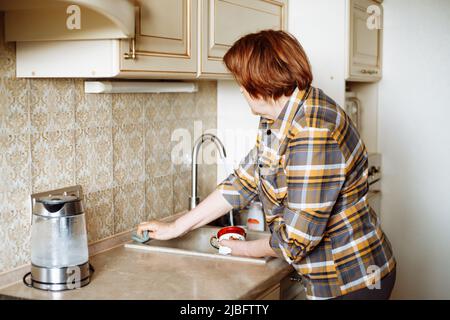 Seitenansicht einer älteren Frau mit dunklem Haar, die in der hellen Küche steht und nach dem Abwaschen das Waschbecken mit einem Schwamm abwischt. Stockfoto