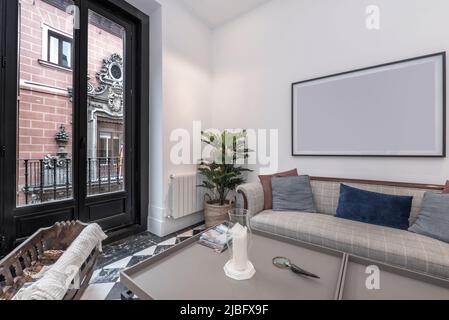 Wohnzimmer mit gepolstertem Sofa mit Holzakken, Marmorböden mit Schachbrettmuster, weißen Sesseln und Balkon mit Aluminiumtüren mit Blick auf ein altes Bui Stockfoto