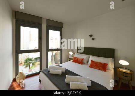 Schlafzimmer mit Queen-Size-Bett mit Kissen und Kissen, sauberen, zusammengerollten Handtüchern und zwei Balkonen mit Blick auf die Straße Stockfoto