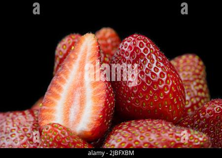 Nahaufnahme von frischen reifen köstlichen roten Erdbeeren ohne grüne Schwänze auf schwarzem Hintergrund. Eine Erdbeere in der Hälfte unter ganze Erdbeeren geschnitten Stockfoto