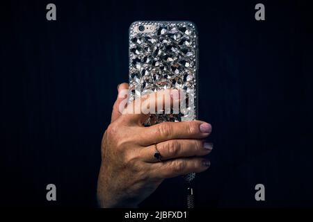 Eine Frauenhand mit einem Ring am Finger hält ein Mobiltelefon in einem mit Strasssteinen auf schwarzem Hintergrund verzierten Etui Stockfoto