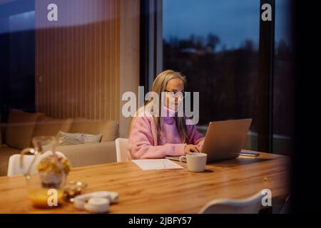 Ältere Frau sitzt drinnen und arbeitet an einem Laptop.Blick durch das Fenster. Stockfoto