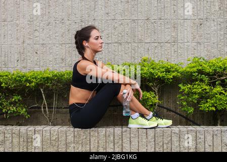 Junge Frau entspannt sich nach dem Sport, sitzt an der grauen Steinmauer, hört Musik, hält eine Flasche Wasser, bereitet sich auf das Laufen vor Stockfoto
