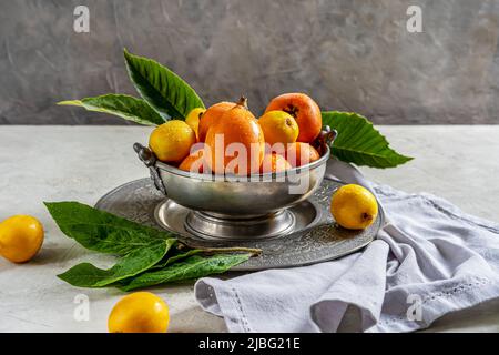 Mispellanobst auf antikem Blechschüssel und Teller mit Blättern auf Betongrund, graue Serviette. Stockfoto