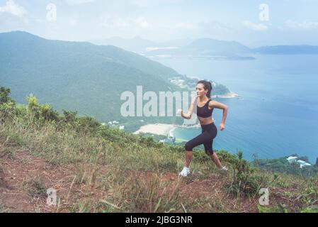 Schlanke junge Sportlerin, die Cardio-Übungen macht, geht den Berg hinauf mit dem Meer im Hintergrund Stockfoto