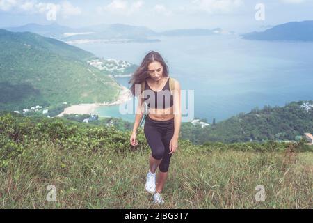 Müde fit Mädchen in schwarzer Sportbekleidung gehen den Hügel mit Bergen und Meer im Hintergrund. Stockfoto