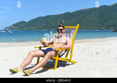 Ein junger glücklicher Mann trinkt Kokosnusssaft, der auf einem Liegestuhl an einem südchinesischen Strand in vietnam sitzt Stockfoto