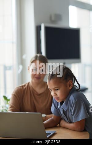 Vertikales Porträt von niedlichen schwarzen Mädchen auf Laptop-Bildschirm mit lächelnden kaukasischen Mutter im Hintergrund Stockfoto
