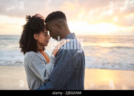 Junges multiethnisches Paar, das bei Sonnenuntergang am Strand in den Armen steht Stockfoto