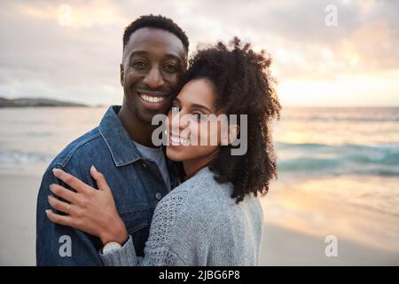 Lächelndes multiethnisches Paar, das Arm in Arm am Strand bei Sonnenuntergang steht Stockfoto