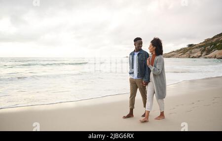 Lächelndes junges multiethnisches Paar, das sich die Hände hält, während es an einem Sandstrand läuft Stockfoto