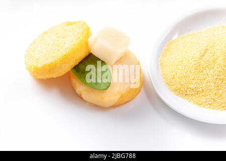 Gebratene Polenta in Form von Pfannkuchen auf weißem Hintergrund mit Käse und Spinat neben einem Teller roher Maiskörner Stockfoto