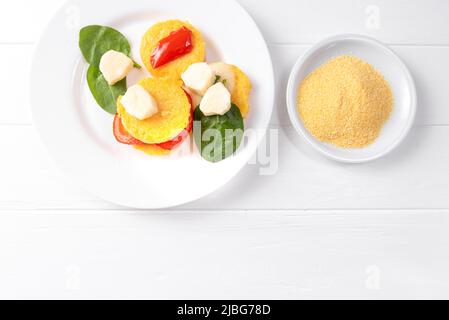 Gebratene Polenta in Form von Pfannkuchen auf weißem Hintergrund mit Käse und Spinat neben einem Teller roher Maiskörner Stockfoto