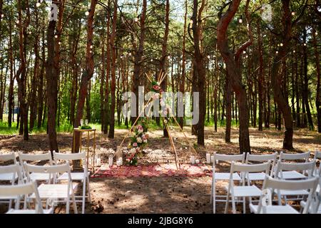 Wunderschöne böhmische Tipi-Bogendekoration auf der Hochzeitsfeier im Freien im Pinienwald mit Kegeln. Stühle, floristische Blumenkompositionen aus Rosen Stockfoto