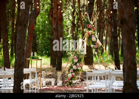 Wunderschöne böhmische Tipi-Bogendekoration auf der Hochzeitsfeier im Freien im Pinienwald mit Kegeln. Stühle, floristische Blumenkompositionen aus Rosen Stockfoto