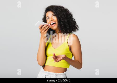 Aufgeregte afroamerikanische Frau, die Sprachnachrichten aufzeichnet, während sie das Smartphone isoliert auf grau hält Stockfoto