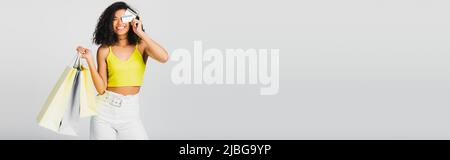 Glückliche afroamerikanische Frau, die Einkaufstaschen hält und das Auge mit Kreditkarten bedeckt, die auf einem grauen Banner isoliert sind Stockfoto
