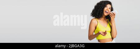 Aufgeregte afroamerikanische Frau, die eine Sprachnachricht aufzeichnet, während sie das Smartphone isoliert auf einem grauen Banner hält Stockfoto