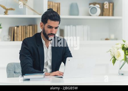 Innenaufnahme eines gut gekleideten kubanischen, hübschen jungen Geschäftsmannes, der an einem Schreibtisch sitzt, auf dem Laptop tippt und konzentriert aussieht. Hochwertige Fotos Stockfoto