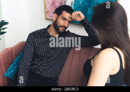 Beunruhigt verärgerten lateinischen bärtigen Mann in seinem 20s sitzt auf einer braunen Couch, gekleidet in einem gemusterten Hemd Blick auf seine hübsche junge kaukasische Freundin. Hochwertige Fotos Stockfoto