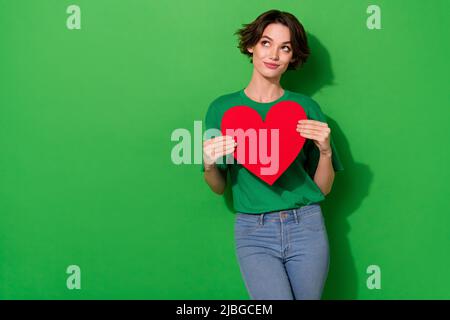 Portrait von minded hübsche Dame halten großes Herz Postkarte suchen leeren Raum auf grünem Hintergrund isoliert Stockfoto