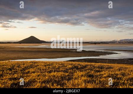 Skútustaðir, Island, 27. April 2022: Blick von einem grasbewachsenen Hügel auf den teilweise mit Eis bedeckten See Myvatn und den Mount Vindbelgjarfjall im goldenen h Stockfoto