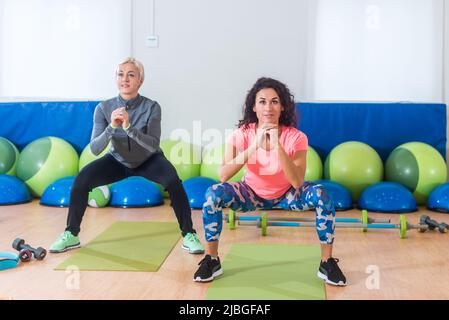 Zwei Frauen in Sportbekleidung hocken beim Training in der Sporthalle mit Körpergewicht. Stockfoto