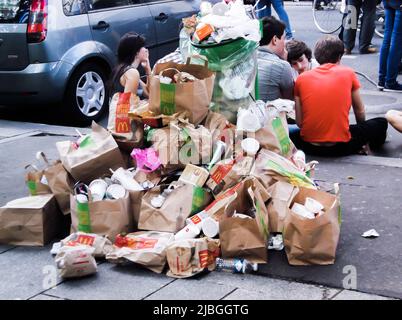 Paris, Frankreich - 11. Juni 2011 : Jugendliche sitzen auf der Straße in der Nähe von McDonald's. Der Mülleimer ist überfüllt mit Müll, Papiertüten und Essensresten Stockfoto