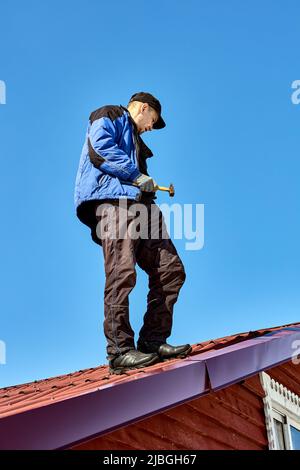 Der Dachdecker steht mit Hammer in der Hand vor dem Hintergrund des blauen Himmels auf dem Dach. Stockfoto