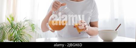 Beschnittene Ansicht einer Frau, die Orangensaft aus dem Krug in das Glas neben der Schüssel, dem Banner, goss Stockfoto