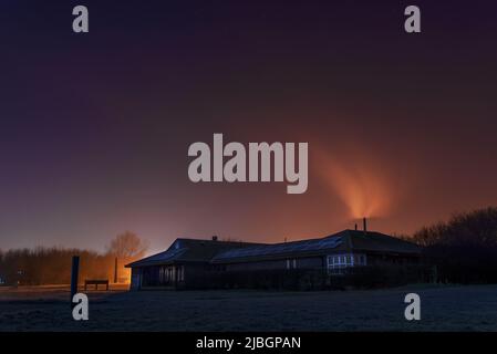 Gebäude in der Ferne mit Rauch aus dem Chimeny. Feld im Vordergrund mit blau und orange Nacht, Abendhimmel und Sterne. Licht hinter dem Haus Stockfoto
