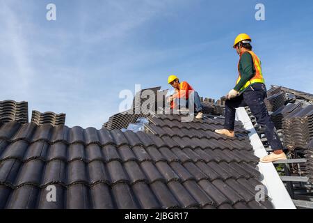 Teamarbeit von 2 Roofern Arbeiter in Schutzuniform tragen und Handschuhe, mit Luft oder pneumatische Nagelpistole und Installation von Beton Dachziegel auf der n Stockfoto