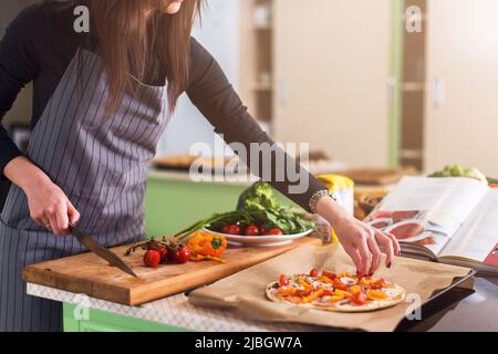 Zugeschnittenes Bild einer Frau, die in der Küche in Scheiben geschnittenes Gemüse auf Pizza-Top-Kochgeschirr legt. Stockfoto