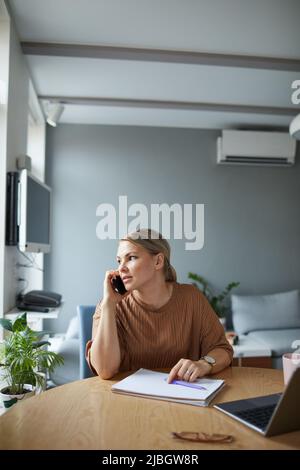 Vertikales Porträt einer jungen kaukasischen Frau, die während der Arbeit oder des Studiums zu Hause in einer Wohnung mit minimalem Dekor telefoniert Stockfoto