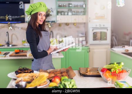 Junge Hausfrau versucht, ein neues Rezept im Kochbuch zu finden, während sie am Tisch mit Essen und Zutaten steht. Stockfoto