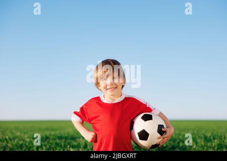 Glückliches Kind gibt vor, ein Fußballspieler zu sein. Porträt eines Kindes im Freien. Junge gegen grünes Feld. Erfolg und Siegerkonzept Stockfoto