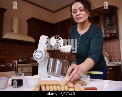Charmante multiethnische Frau, Konditorei, Hausfrau bereitet traditionelle italienische Dessert Tiramisu Kuchen mit Schlagsahne, Mascarpone-Käse und Dame Stockfoto
